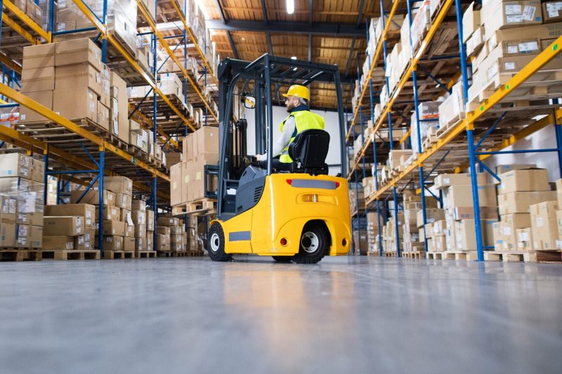 Young male worker lowering a pallet with boxes. Forklift driver working in a warehouse.